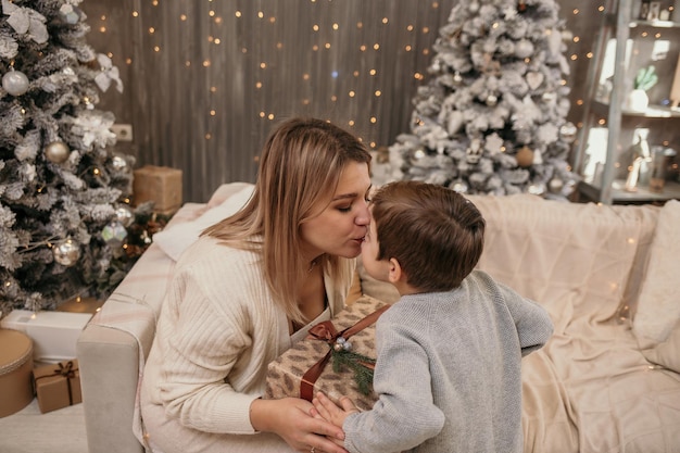 Mom is sitting on the couch and kissing her son in the room near the Christmas tree