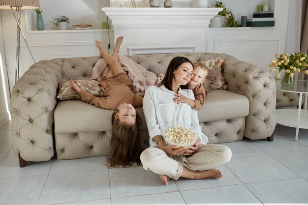 Mom and kids eat popcorn at home on a day off. A woman a boy and a girl relax on the couch and hug