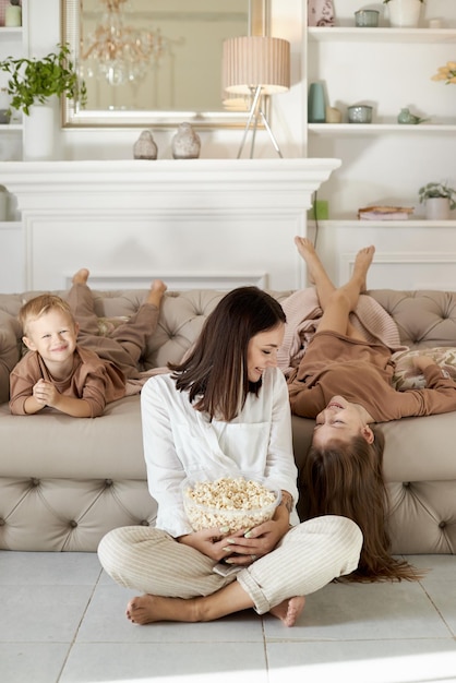 Mom and kids eat popcorn at home on a day off. A woman a boy and a girl relax on the couch and hug