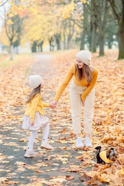 La mamma con un cappello a maglia guarda delicatamente sua figlia di cinque anni tenendole la mano in autunno sullo sfondo di foglie gialle cadute, accanto al loro animale domestico un piccolo cane