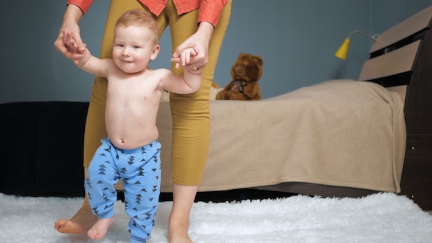 Mom leads baby holding his hands forward to camera. First baby steps concept. Medium shot