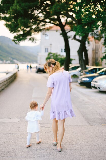 Mom leads her little daughter by the hand along the embankment of perast city past cars and trees