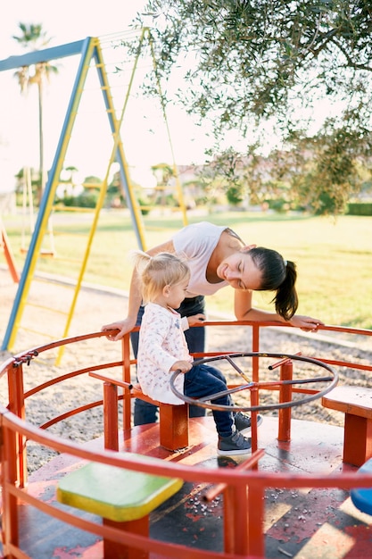 Photo mom leaned over to a little girl sitting on a carousel on the playground