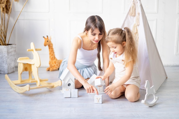 Mom and little baby daughter are playing at home in a bright children's room among wooden toys smiling and having fun happy motherhood