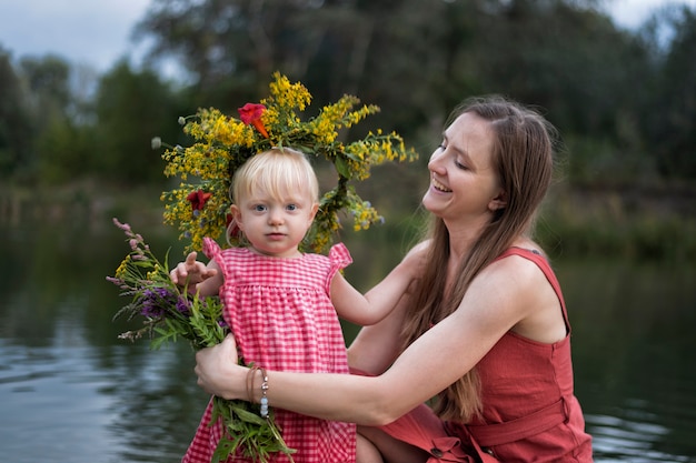 Mom and little daughter are resting by the river. Child in wreath of flowers.