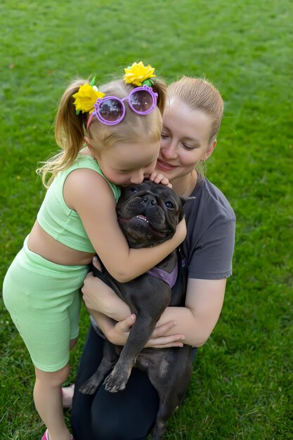 Mom and little daughter sit on the lawn in the park and hug the dog