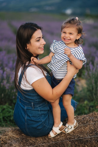 Mom and little daughter sits on hay by the farm Background of summer lavender field Family denim style Little girl hug mom