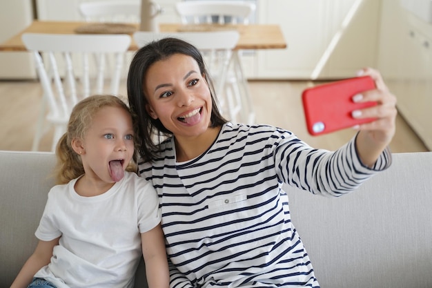 Mom little daughter take selfie photo by phone show tongues together having fun sitting on sofa
