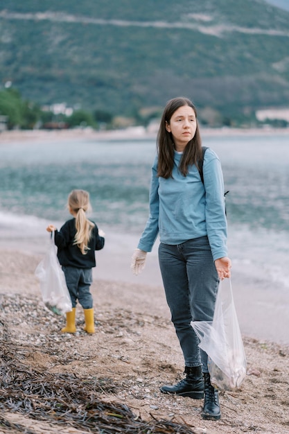 Mom and little girl collect garbage in bags while standing on the beach by the sea