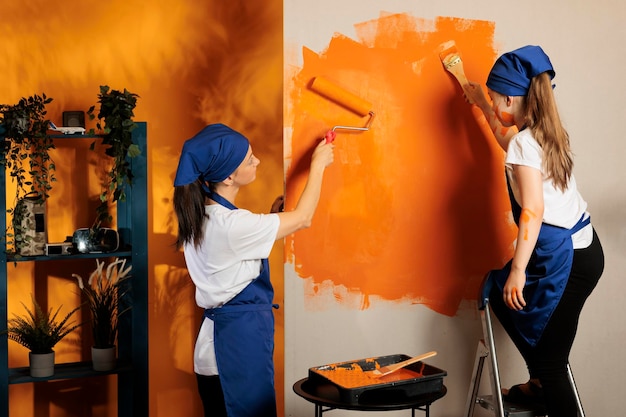 Mom and little girl painting room walls, sitting on ladder and\
using painting roller tool or paintbrush with bristles. happy\
people wokring on house decor improvement with decoration\
roll.