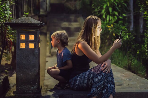 Mom looks into her smartphone, the son looks into the luminous cozy window of the house. the boy lacks attention, care, love and home comfort