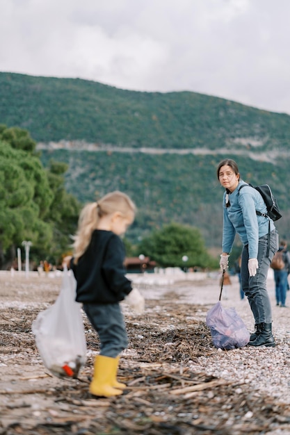 Mom looks at a little girl helping her collect garbage on the beach into bags