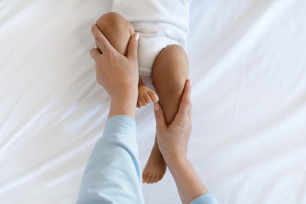 Mom Making Development Gymnastics With Black Baby Lying On Bed At Home