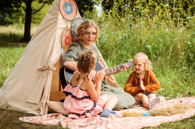 Mom plays the guitar to her children, little daughters. Multicultural festival or children's party. Family are sitting next to wigwam or teepee decoration. Boho style decorations.