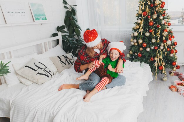 Mom plays with her son on the bed at home near the christmas tree. woman and child in santa claus hats. Christmas.