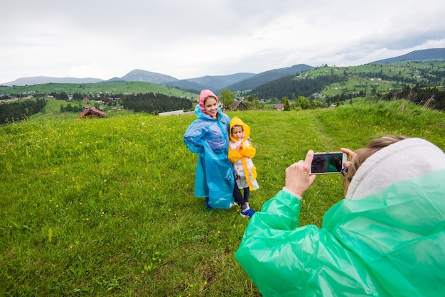 Мама в плаще фотографирует двух своих симпатичных детей в плащах на фоне живописных лугов.