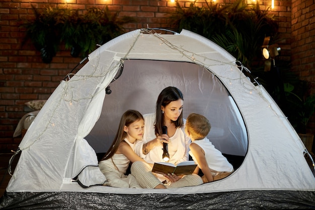 Mom reads children a bedtime story sitting in a tent at home. Mother son and daughter hug and read a book with a flashlight in their hands