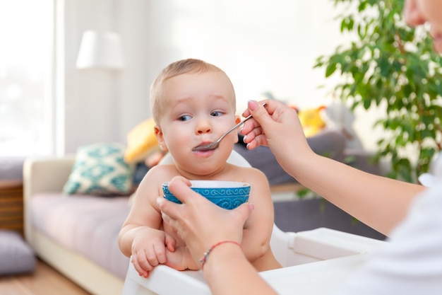 Mom's hand feeds funny cute happy blond baby toddler with spoon from blue bowl, apartment background, copy space, horizontal