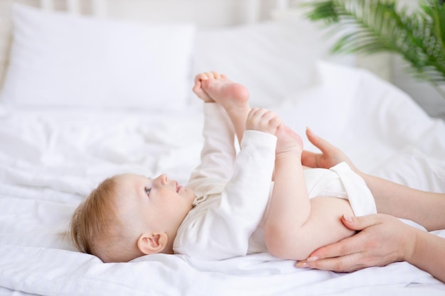 Mom's handen houden de benen baby 6 maanden oude blonde jongen op een wit bed in een lichte slaapkamer na het slapen in de ochtend in een katoenen bodysuit het concept van kinderartikelen