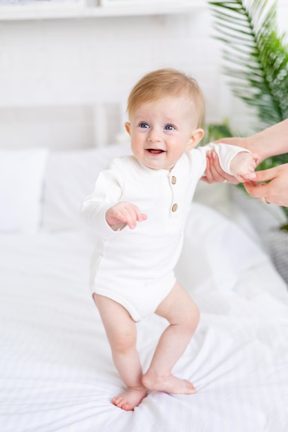 Mom's handen houden een baby van 6 maanden oud een blonde jongen met blauwe ogen op een wit bed in een lichte slaapkamer in een witte katoenen bodysuit het concept van kinderartikelen