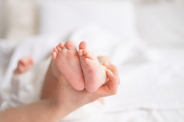 엄마의 손은 아기 용품과 액세서리, 엄마의 사랑과 보살핌이라는 개념의 흰색 면 침구가 있는 집에 있는 흰색 침대에 초점을 맞춘 아기의 발을 잡고 있습니다.