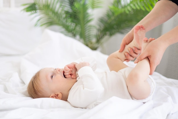 Mom's hands hold the legs baby 6 months old blond boy on a white bed in a bright bedroom after sleeping in the morning in a cotton bodysuit the concept of children's goods