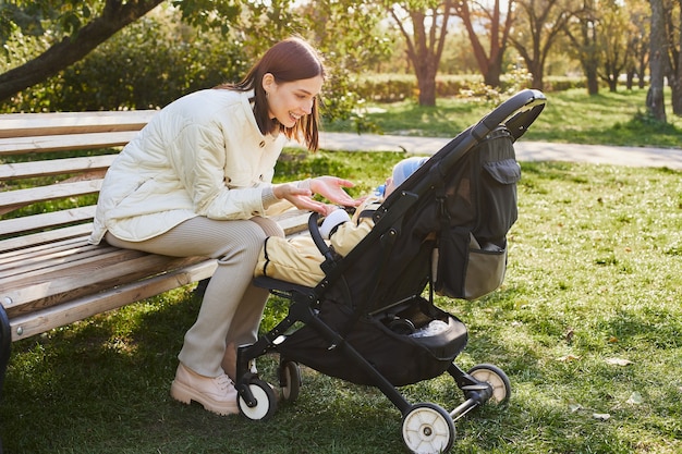 Mom sits on a bench near the stroller with her little son in the park.