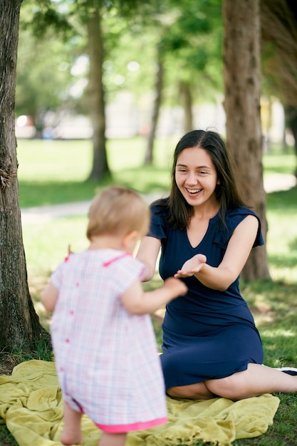 Mom sits on a blanket on the lawn and stretches out her arms to a little girl walking towards her