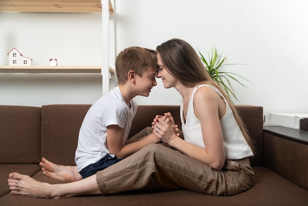 Mom and son are sitting on couch with closed eyes and foreheads leaning against each other