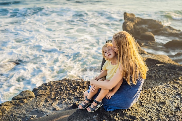 엄마와 아들은 바위에 앉아 바다를 바라보고 있습니다. 세로 여행 관광객 - 아이와 엄마. 긍정적인 인간의 감정, 활동적인 생활 방식. 바다 해변에서 행복 한 젊은 가족