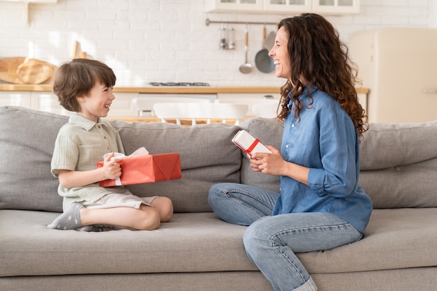 Mom and son celebrate birthday together happy mum and excited son sit on couch hold gifts smiling