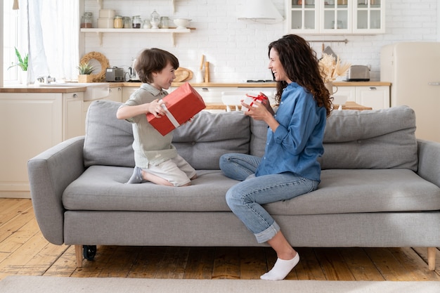 Mom and son celebrate birthday together happy mum and excited son sit on couch hold gifts smiling