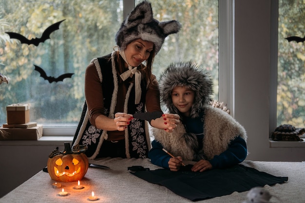 Photo mom and son in halloween costumes play together and make paper bat halloween decorations on pumpkins