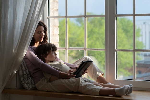 Mom and son kid sit on windowsill look at tablet screen mother or nanny spend time with son at home