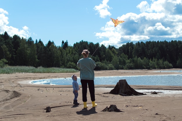 Mom and son play flying kite on river shore