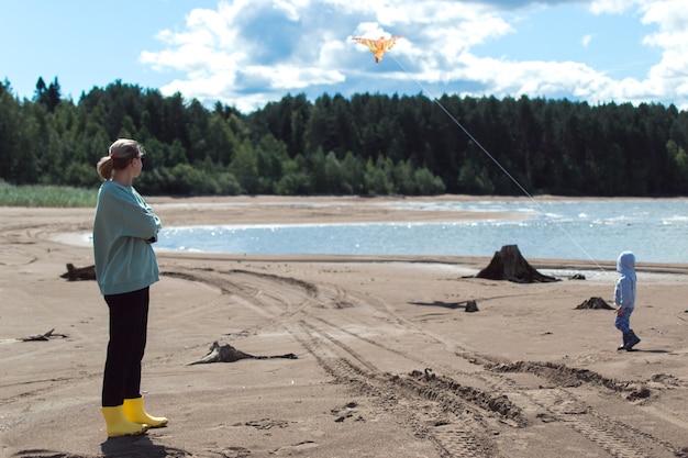 Mom and son play flying kite on river shore