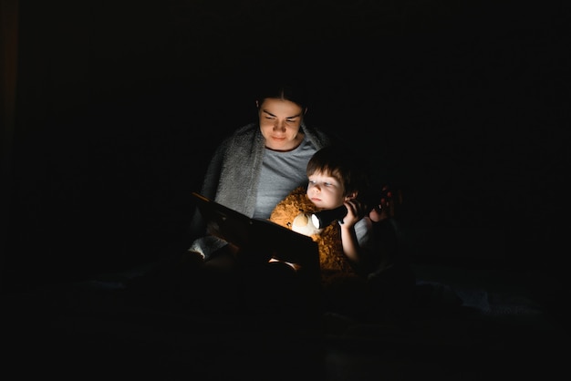 담요 아래에 손전등으로 책을 읽고 엄마와 아들