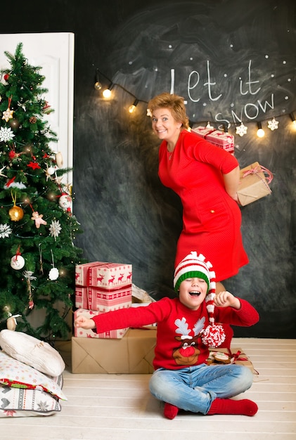 Mom and son in red Christmas sweaters, having fun at the Christmas tree, hugging and laughing