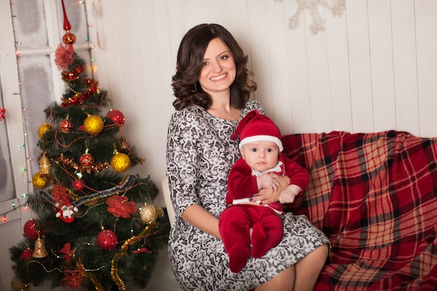 Mom and son in Santa Claus costume in a home setting near a Christmas tree against a white wooden wall