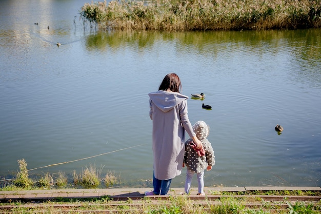 Mom stands with her little daughter on the shore of the lake in autumn clothes