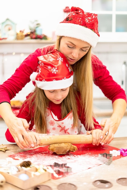 Мама учит дочку раскатывать тесто для печенья на кухне в новогодней шапке
