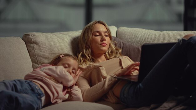 Mom using laptop next to daughter on couch Girl falling asleep mom shoulder