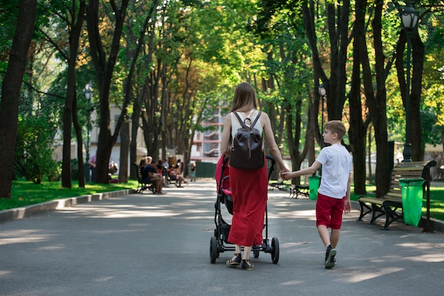 Мама гуляет в парке с коляской и старшим сыном. Прогулка с детьми. Детский образ жизни