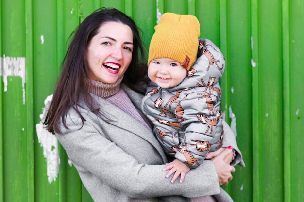 Mom with a baby on a green industrial door. in a warm gray\
coat. portrait of a beautiful girl with her son on a green\
industrial door. holds a one-year-old child in his arms and smiles,\
the child also laughs