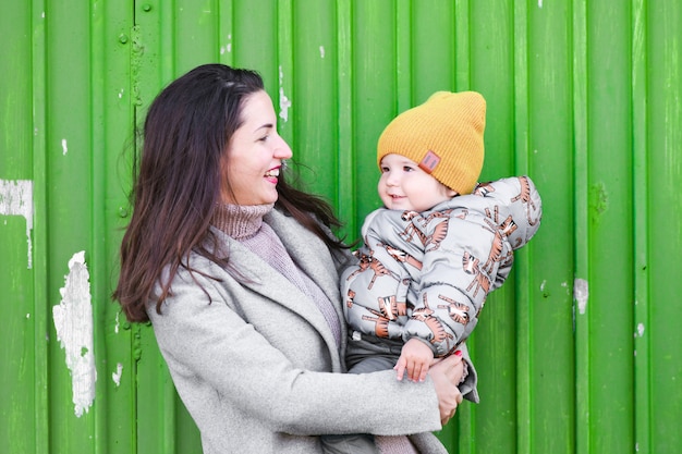 Mom with a baby on a green industrial door. in a warm gray\
coat. portrait of a beautiful girl with her son on a green\
industrial door. holds a one-year-old child in his arms and smiles,\
the child also laughs