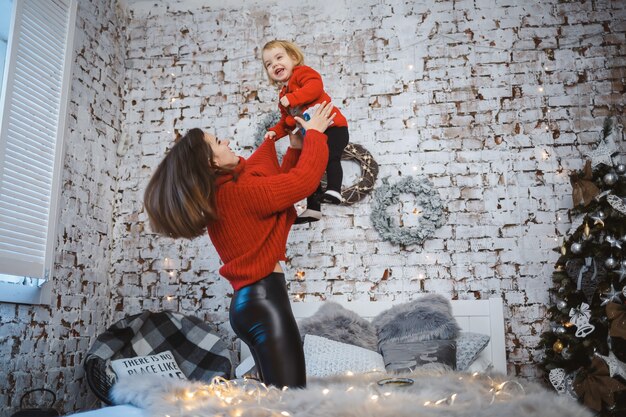 Mom with daughter in red warm sweaters are jumping on the bed. Happy motherhood. Warm family relationships. Christmas and New Year's interior. Love. Family concept.