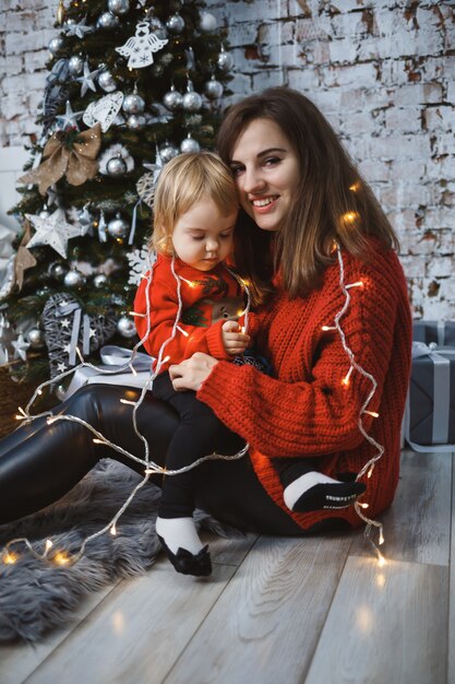 Mom with daughter in red warm sweaters are jumping on the bed. Happy motherhood. Warm family relationships. Christmas and New Year's interior. Love. Family concept.