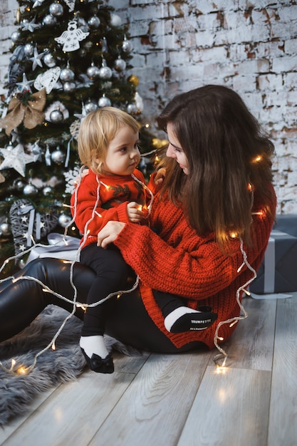 Mom with daughter in red warm sweaters are jumping on the bed. Happy motherhood. Warm family relationships. Christmas and New Year's interior. Love. Family concept.