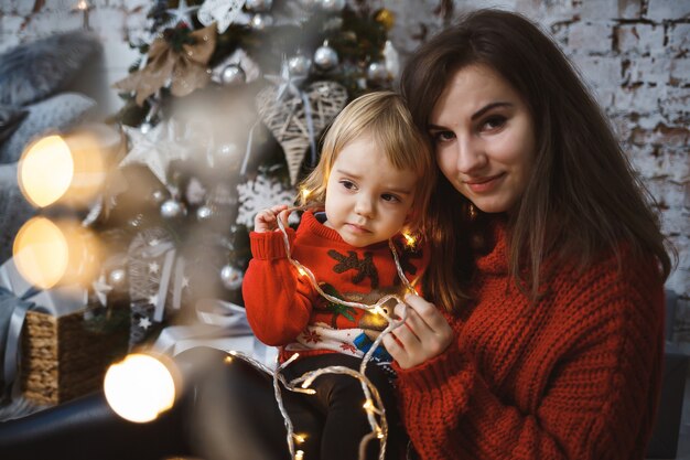 赤い暖かいセーターを着た娘を持つお母さんがベッドの上でジャンプしています。幸せな母性。暖かい家族関係。クリスマスとお正月のインテリア。愛。家族の概念。