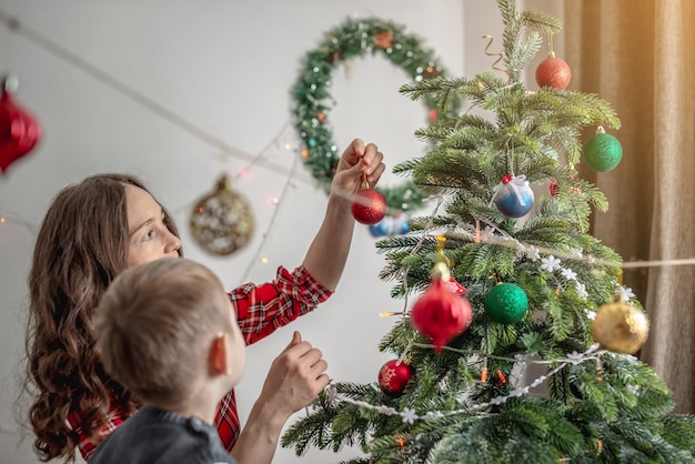 그녀의 아이와 엄마는 장난감과 공으로 크리스마스 트리를 함께 장식하고 있습니다. 새해, 아늑한 분위기 및 축제 분위기를 위한 가족 준비의 개념.